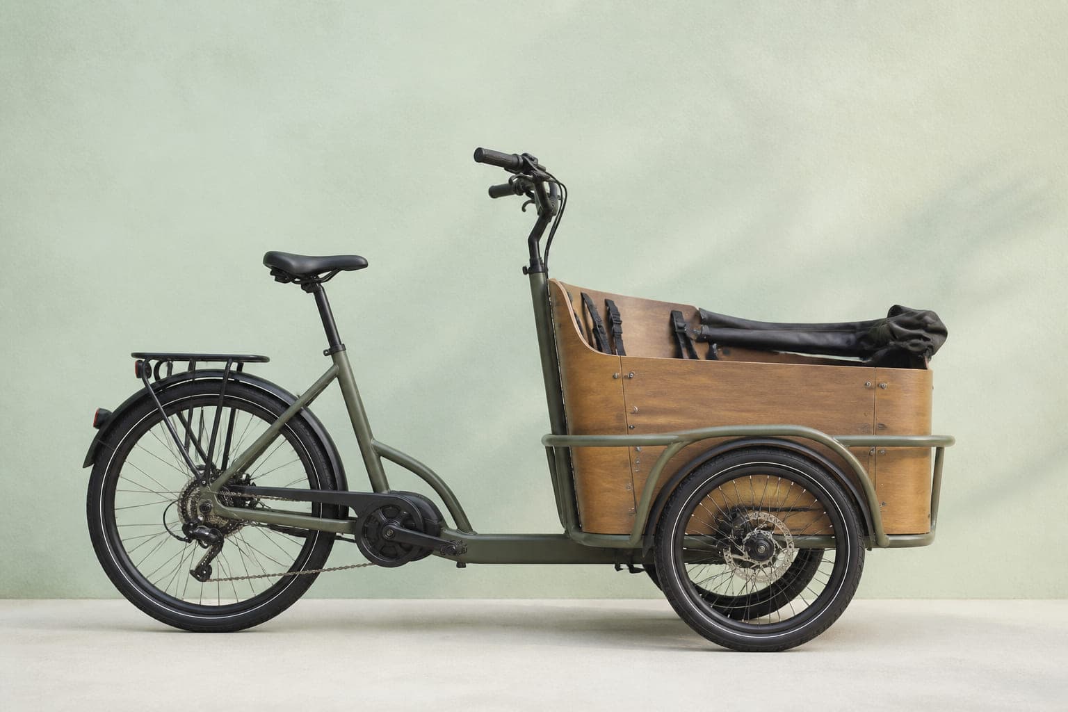 Dreirädriges Lastenrad in Olivgrau mit großem Holzkasten zwischen zwei Vorderrädern und zwei Gurten darin, von der Seite